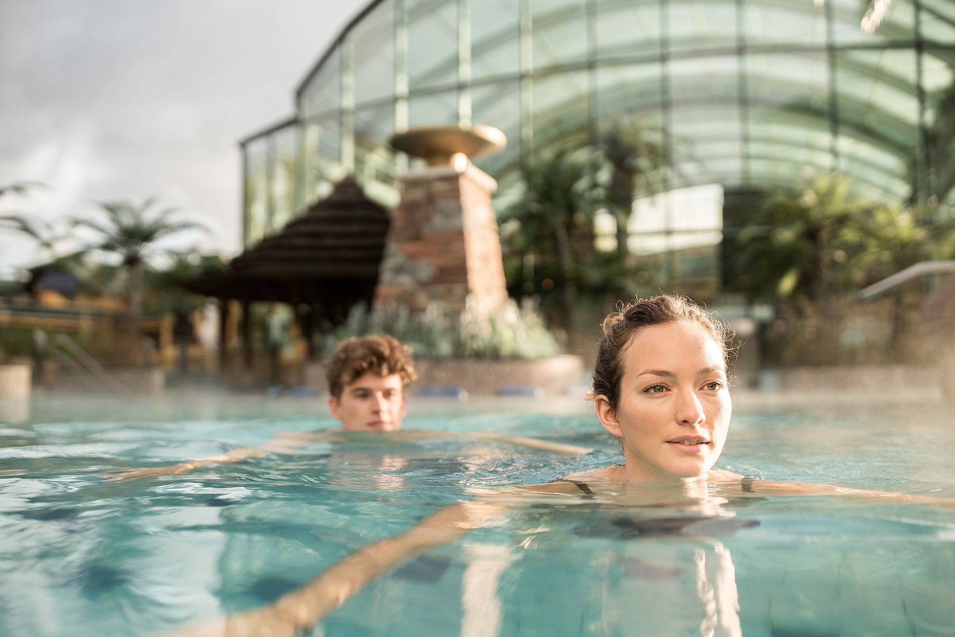 Eine Frau und ein Mann schwimmen hintereinander im Wasser des Au&szlig;enbeckens des EurothermenResorts Bad Schallerbach, im Hintergrund unscharf das gl&auml;serne Cabrio-Dach der Therme Tropicana.